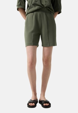 Osoba ubrana w oliwkowozielone szorty z elastycznym pasem, zestawione z czarnymi sandałami na krzyżujące się paski na jednolitym tle.