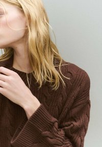 Brauner Kabelstrickpullover mit weiten Ärmeln und strukturierten Mustern. Modell teilweise sichtbar, mit zerzausten blonden Haaren und einer Hand nahe am Kragen.