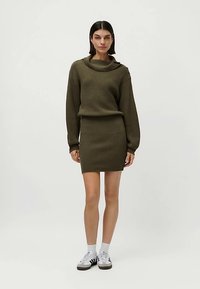 Olivenfarbener Strickpullover und Rock im Set. Der Pullover hat einen Wasserfallausschnitt und eine gerippte Struktur; der Rock ist eng anliegend. Kombiniert mit weißen Sportsocken und Turnschuhen.