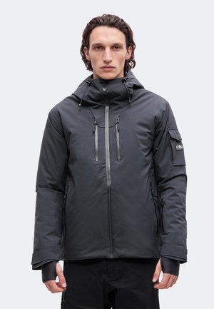 Un homme porte une veste à capuche gris foncé avec une fermeture éclair sur le devant, des fermetures éclair sur la poitrine et une poche sur la manche, sur un fond clair uni.