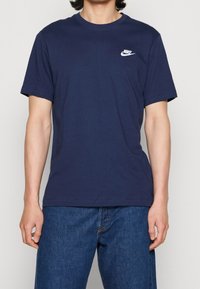 Laivansininen puuvillapaita, jossa on pyöreä pääntie, lyhyet hihat ja valkoinen Nike-logo vasemmassa rinnassa. Sujuva tekstuuri ja rento istuvuus.