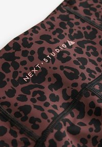 Leggings à imprimé léopard dans des tons de brun foncé et de noir, avec une texture lisse, des coutures détaillées, et un logo "NEXT STUDIO" rose.