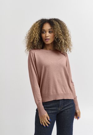 Pull en maille rose clair avec une coupe décontractée, poignets et ourlet côtelés, associé à un jean en denim bleu foncé. Tissu doux et texturé.