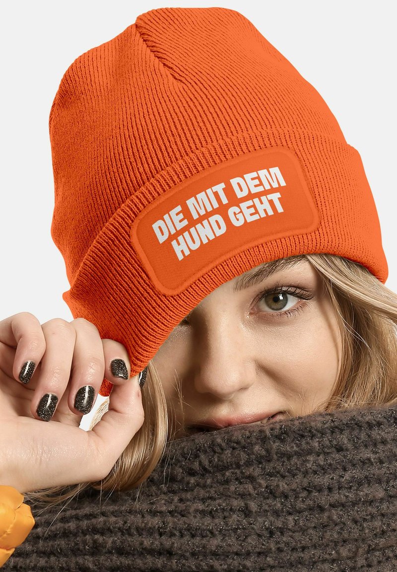 Bright orange knitted beanie with a ribbed texture and a rectangular patch featuring white text that reads "DIE MIT DEM HUND GEHT."