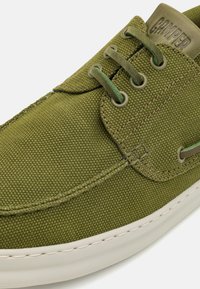 Green canvas sneaker featuring a rounded toe, lacing system, and embossed logo on the tongue. White rubber sole with stitching details.