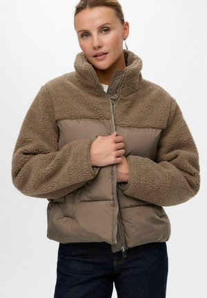 Ženska, oblečena v rjavo dvobarvno flis jakno z vzorcem šivanja, jo zateguje z zadrgo, oblečena v temne hlače, na enostavni svetli podlagi.