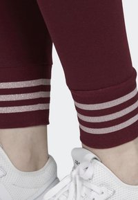 Burgundowe legginsy sportowe z szerokimi, srebrnymi paskami brokatowymi przy mankietach. Teksturowana tkanina i dopasowany krój. Uzupełnione białymi sneakersami.