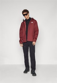 Chaqueta impermeable roja con capucha, llevada sobre un suéter negro y pantalones negros. Unas gafas de sol negras y botas de senderismo completan el conjunto.