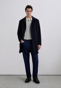 Abrigo de lana azul marino con cierre de botones, llevado sobre un polo con rayas crema y azul marino, combinado con pantalones de mezclilla oscura y zapatos negros.