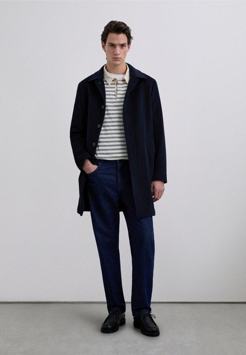 Abrigo de lana azul marino con cierre de botones, llevado sobre un polo con rayas crema y azul marino, combinado con pantalones de mezclilla oscura y zapatos negros.
