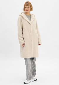 Crème kleurige, fluffy, knielange jas met een capuchon en lange mouwen. Draag boven een grijze top en zilver glanzende broek, met een eenvoudig ontwerp.
