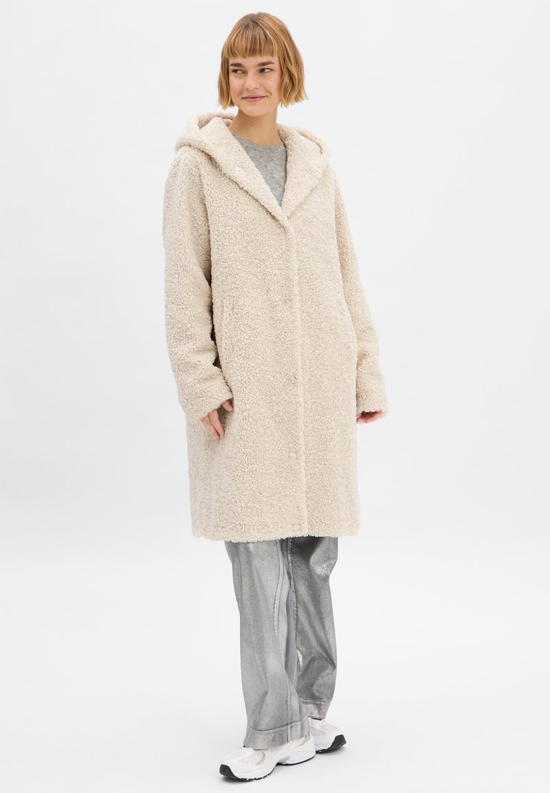 Crème kleurige, fluffy, knielange jas met een capuchon en lange mouwen. Draag boven een grijze top en zilver glanzende broek, met een eenvoudig ontwerp.