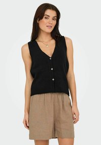 Top sin mangas negro con escote en V y tres botones en crema. Llevado sobre pantalones cortos sueltos de color marrón claro con un acabado texturizado.