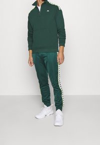 Sweatshirt zippé vert avec un col haut, présentant des accents jaunes. Joggers verts assortis avec des bandes latérales et des baskets blanches.
