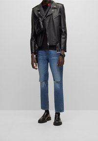 Schwarze Lederbikerjacke mit Reißverschlüssen, eng anliegendes schwarzes Hemd, zerrissene blaue Jeans und klobige schwarze Loafers. Kleine schwarze Umhängetasche.