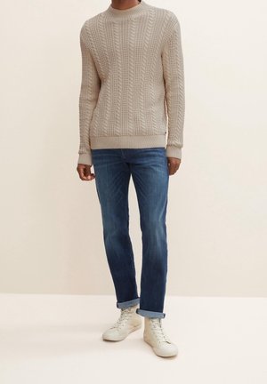 Homme portant un pull beige en maille torsadée, un jean bleu retroussé et des baskets montantes blanches, debout devant un fond uni.