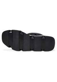 Semelle de sandale noire avec grip texturé et segments de bande de roulement surélevés, vue de dessous sur fond blanc.