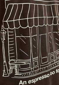 Brown T-shirt featuring a white line drawing of a café storefront with a striped awning, windows, a table, and a chair beside a streetlamp.