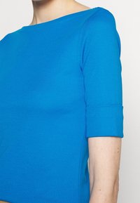 Top azul de manga corta hecho de una tela suave y elástica. Cuello redondeado, corte entallado y detalles de costura visibles en las mangas.