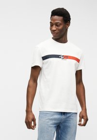 Άνδρας που φοράει λευκό μπλουζάκι Tommy Jeans με ριγέ σε ναυτικό μπλε και κόκκινο, συνδυασμένο με ανοιχτόχρωμα τζιν, στέκεται μπροστά σε απλό φωτεινό φόντο.