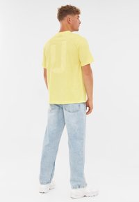 Żółta bawełniana koszulka z krótkim rękawem, z dużym, jasnym logo "J" z tyłu. Połączona z jasnoniebieskimi dżinsami.