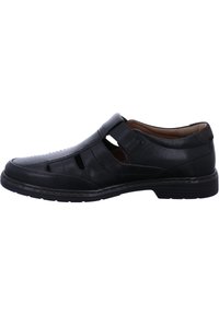 Black leather shoe with open cutouts, flat sole, and a Velcro strap. Features smooth texture and stitching accents. Designed for comfort and breathability.