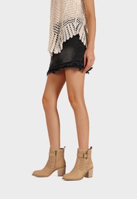 Stivaletti alla caviglia in suede color marrone chiaro con motivi intagliati e cerniera laterale. Il design presenta un tacco basso a blocco e una fibbia decorativa.