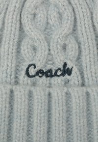 Beanie lavorato a maglia di colore azzurro chiaro con motivo a treccia, caratterizzato da un logo "Coach" ricamato in blu scuro sul davanti e una fascia a coste nella parte inferiore.