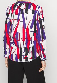 Långärmad skjorta tillverkad av lättviktig tyg som har en vit botten med djärva röda, lila och svarta abstrakta geometriska mönster.