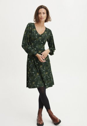 Vihreä kukkakuosiset mekko, jossa on V-aukko, pitkät puffihihat ja levenevä helma. Yhdistetty mustien sukkahousujen ja ruskeiden nilkkureiden kanssa.