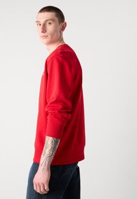 Rød sweatshirt med lange ærmer og ribbede manchetter, med en rund halsudskæring. Båret med mørk denimjeans, som viser en synlig tatovering på armen.