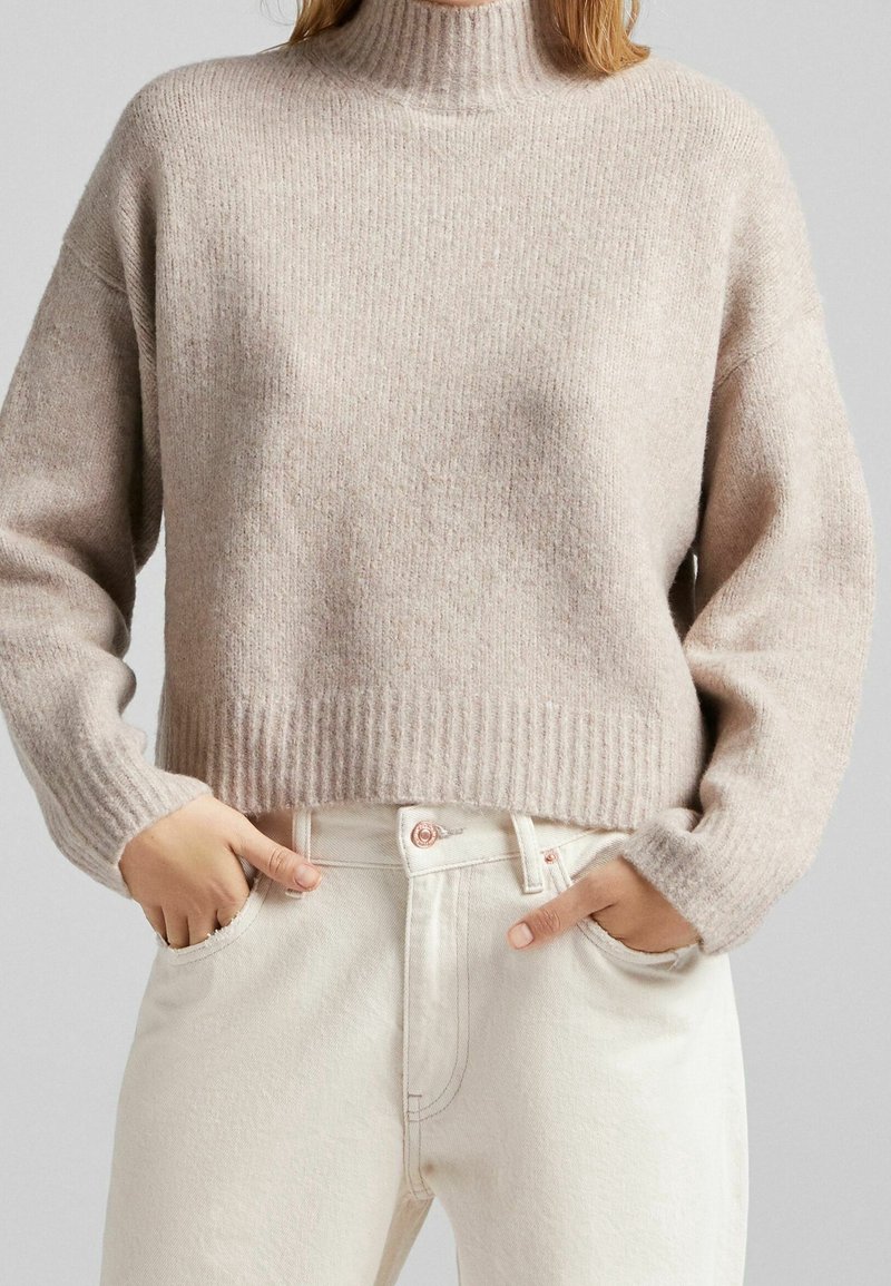 Beige geribbelde gebreide trui met een hoge hals en verlaagde schouders, gecombineerd met lichte denim met een subtiele roze knoopdetail.