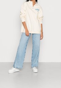 Vit zip-up sweatshirt med blå "NIKE" logotyp, kombinerad med ljusblå vida jeans och vita sneakers på ett ljusgrått golv.