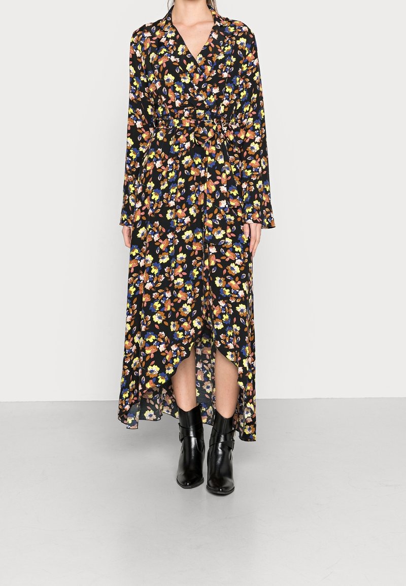 Femme portant une robe portefeuille à manches longues noire à motifs floraux avec un ourlet asymétrique et des bottines noires, debout devant un fond uni.