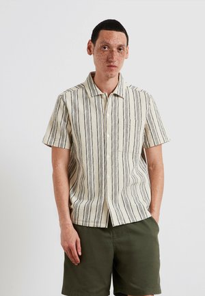 Young man with short dark hair and freckles wearing a beige striped short-sleeve button-up shirt and olive green shorts, standing relaxed.