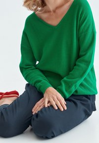 Pull en maille côtelé vert, avec un large col en V, coupe ample et manches longues, porté avec un pantalon foncé à coupe décontractée et des sandales à lanières rouges.