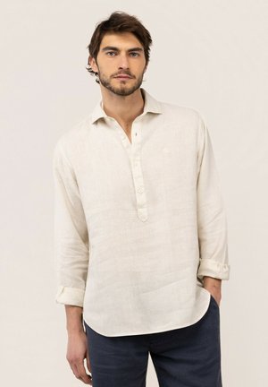 Hombre con cabello oscuro y barba lleva una camisa de lino color marfil con botones y pantalones oscuros, de pie frente a un fondo claro y liso.