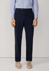 Uomo che indossa pantaloni blu navy testurizzati, una camicia a maniche lunghe a righe blu chiaro e scarpe in suede beige con lacci, in piedi contro uno sfondo neutro.