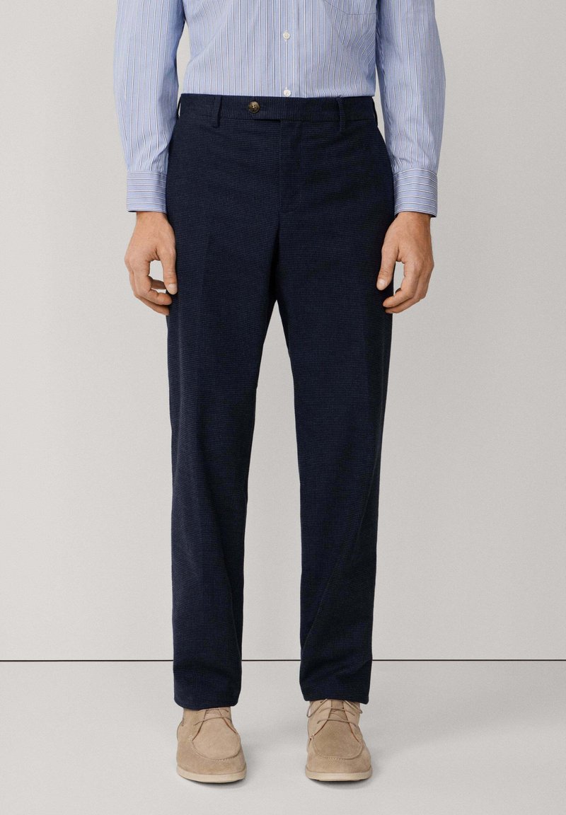Uomo che indossa pantaloni blu navy testurizzati, una camicia a maniche lunghe a righe blu chiaro e scarpe in suede beige con lacci, in piedi contro uno sfondo neutro.