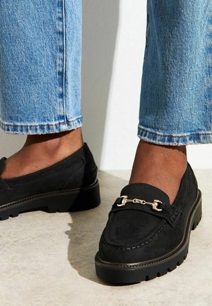 Black suede loafers with a gold chain accent, featuring a thick rubber sole and rounded toe. Styled with light blue cropped jeans.