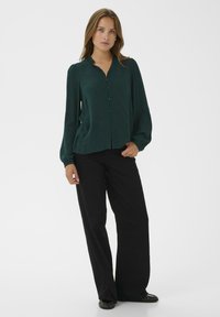 Donkergroene blouse met lange mouwen, een gerimpelde kraag en knoopsluiting aan de voorkant, gecombineerd met zwarte wijde broek en zwarte platte schoenen.