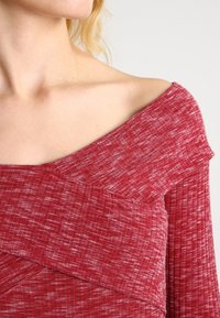 Top rosso a spalle scoperte con tessuto testurizzato e design a strati. Il materiale presenta un delicato motivo melangiato, che aggiunge profondità al colore.