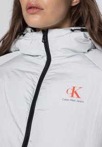 Světle šedá bunda na zip s vysokým límcem, černým zipem a logem Calvin Klein Jeans v oranžové barvě. Tkanina má hladký, lesklý povrch.