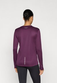 Pitkähihainen urheilupaita syvän violetista kankaasta, jossa on pyöreä kaula-aukko, resoitettu pinta ja heijastavat yksityiskohdat takareunassa.