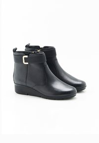 Botines negros con acabado de cuero suave, banda elástica en la parte superior, cremallera lateral y un tacón bajo en cuña, con un acento dorado.