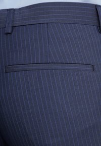 Detailní záběr na zadní kapsu kalhot tmavě modrého pruhovaného obleku s odpovídajícím poutkem na opasek a vodorovným otvorem kapsy.