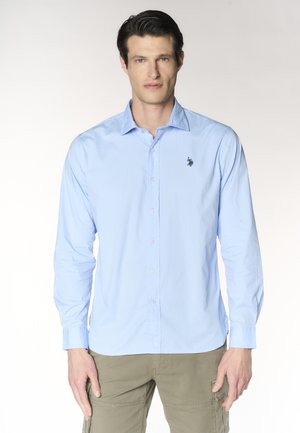 Uomo con camicia azzurro chiaro abbottonata con piccolo logo ricamato sul petto e pantaloni cargo color kaki, in piedi rivolto in avanti.