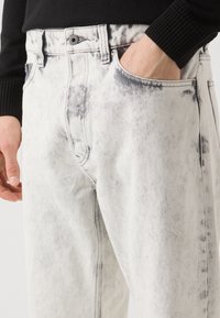 Baltas denim bikses ar izbalējušu melnu rakstu, piecu kabatu dizains un kontrastējošas šuves. Audumam ir gluda tekstūra.