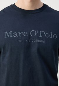 Tumši zila kokvilnas T-kreklu ar apaļu izgriezumu, kurā uz krūtīm ir balts teksts "Marc O'Polo EST. IN STOCKHOLM".