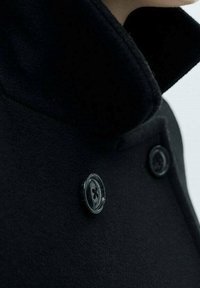 Abrigo de lana negra con un cuello alto, que cuenta con dos botones claros y una textura suave. El diseño es estructurado y ajustado.